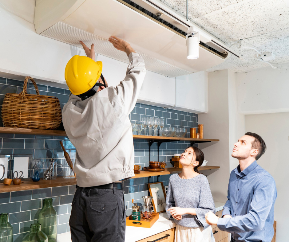 A technician wearing a yellow hard hat and light-colored work jacket is inspecting or repairing a wall-mounted air conditioning unit in a modern kitchen. A technician wearing a yellow hard hat and light-colored work jacket is inspecting or repairing a wall-mounted air conditioning unit in a modern kitchen.